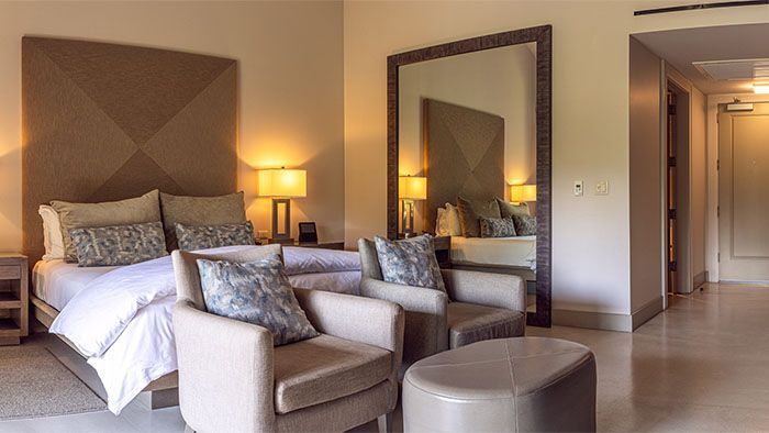 Luxurious bedroom with bed, two armchairs, large mirror, warm lighting.