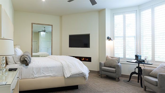 Bedroom with bed, fireplace, TV, chairs, window with shutters; neutral colors.