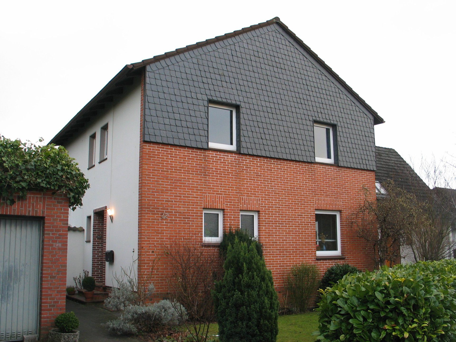 Zweistöckiges Haus mit Backsteinfassade im unteren Bereich und dunkelgrauer Holzverkleidung im oberen Bereich.