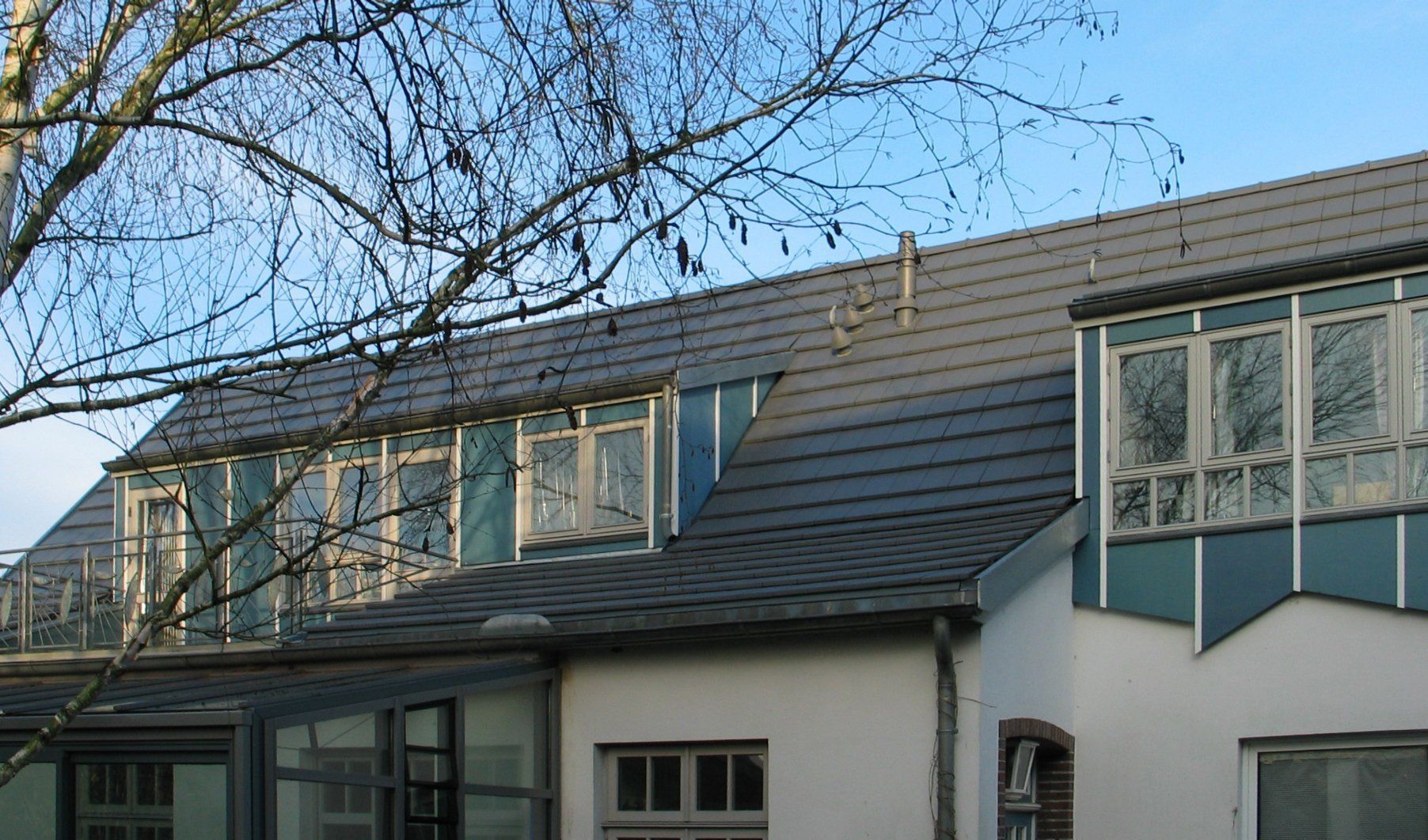 Ein Gebäude mit grauem Ziegeldach, blauen Fensterrahmen und kahlen Ästen vor blauem Himmel.