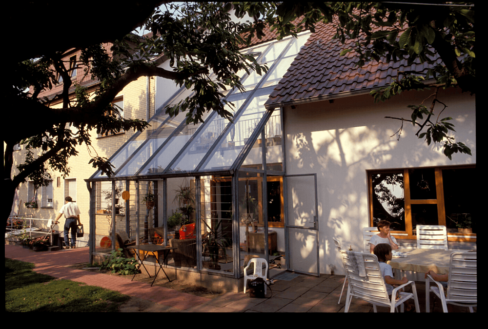 Wintergartenanbau an ein Haus mit Tisch und Stühlen im Außenbereich. Personen sind zu sehen, Bäume im Vordergrund.