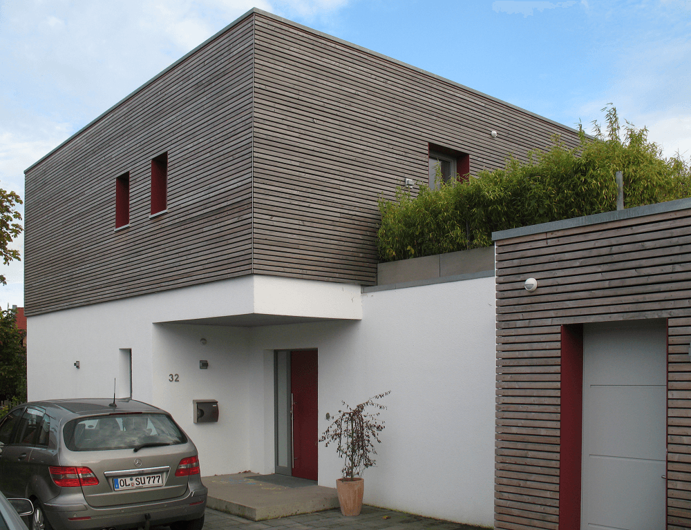 Modernes Haus mit weißem Erdgeschoss, hölzernem Obergeschoss und roter Tür. Ein Auto ist in der Nähe geparkt.