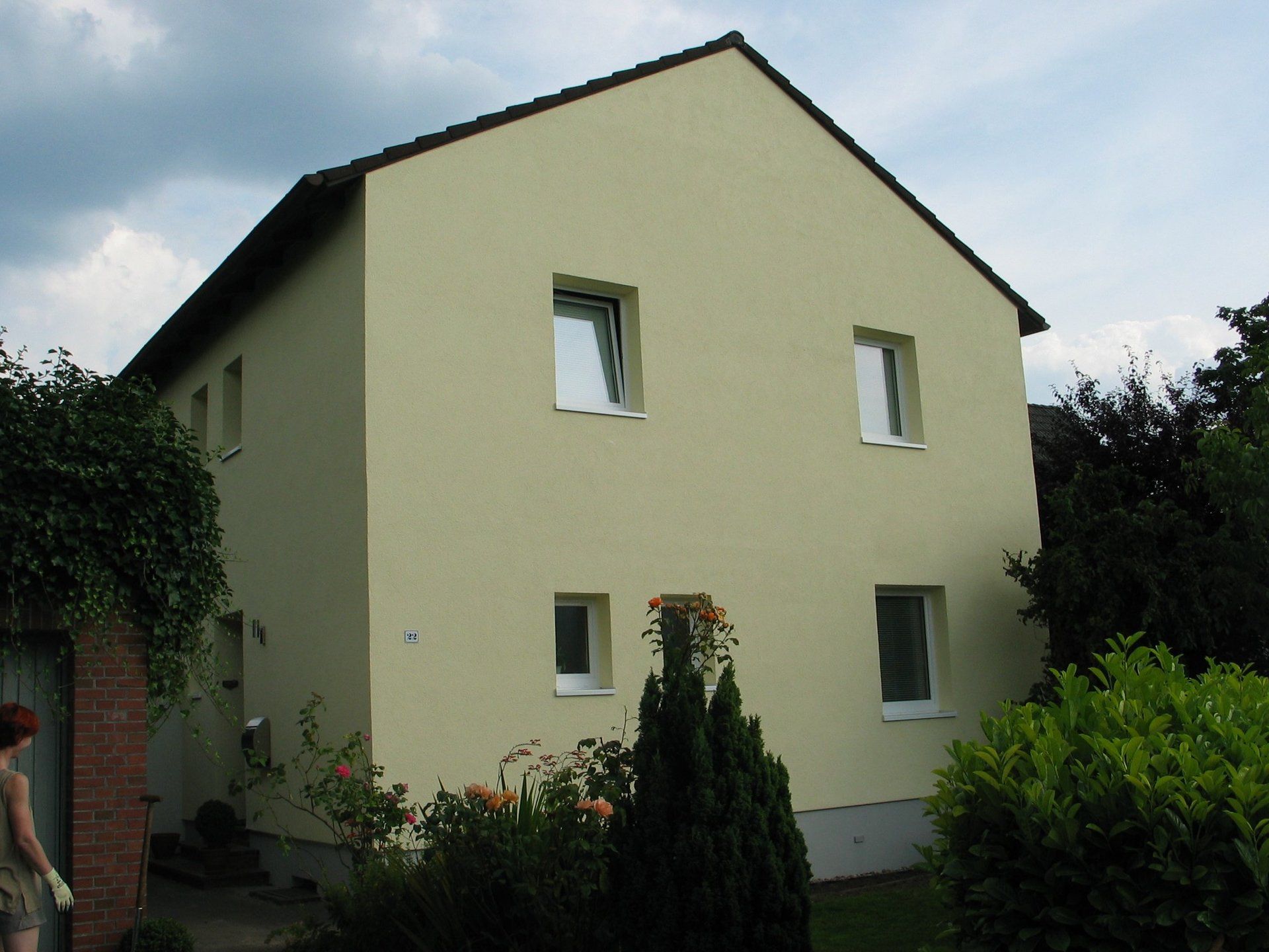 Zweistöckiges beigefarbenes Haus mit weiß gerahmten Fenstern, umgeben von Grün und einer Backsteinmauer.