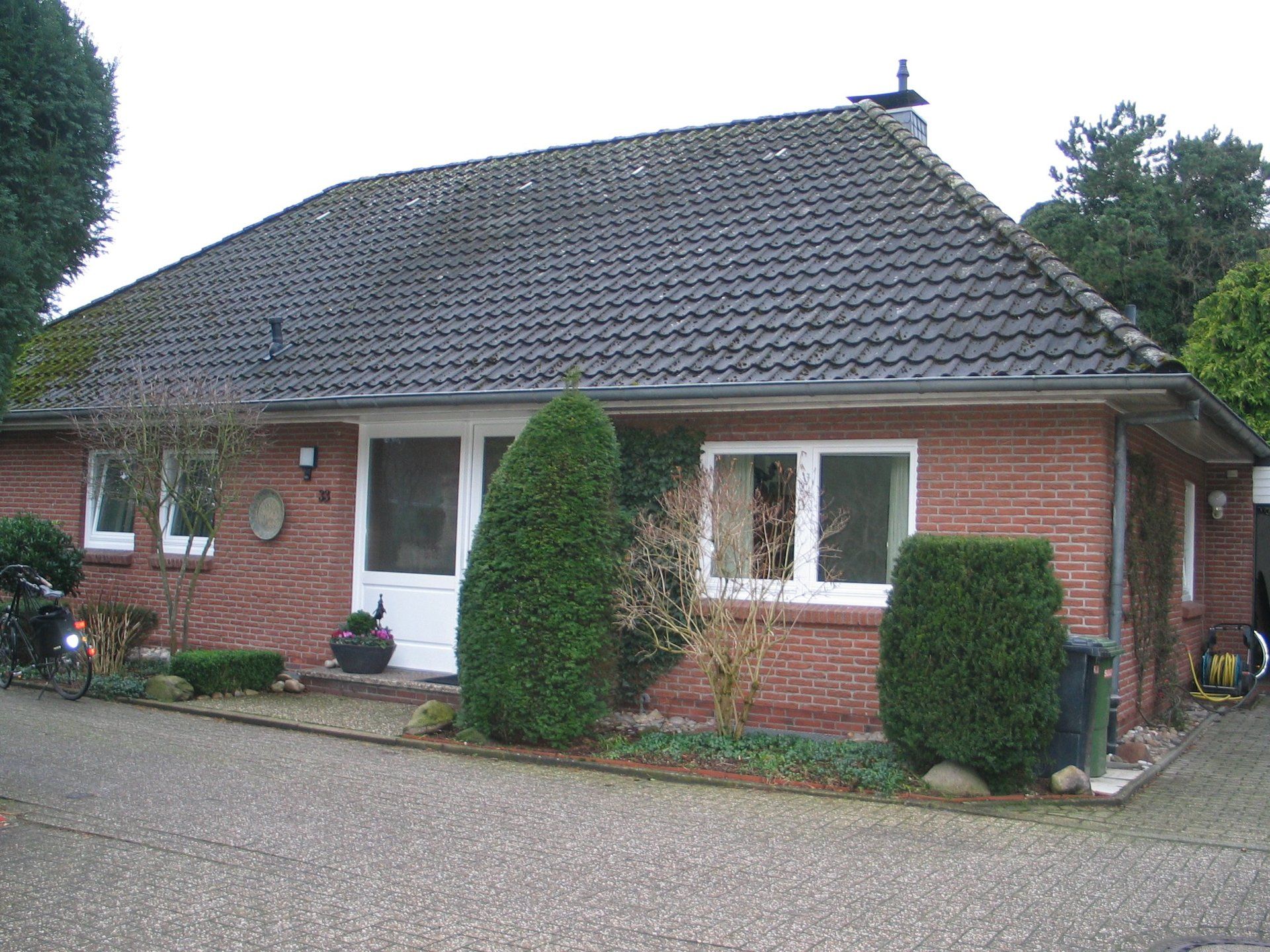 Backsteinhaus mit dunklem Ziegeldach, weißen Fenstern und Tür sowie Gartengestaltung.