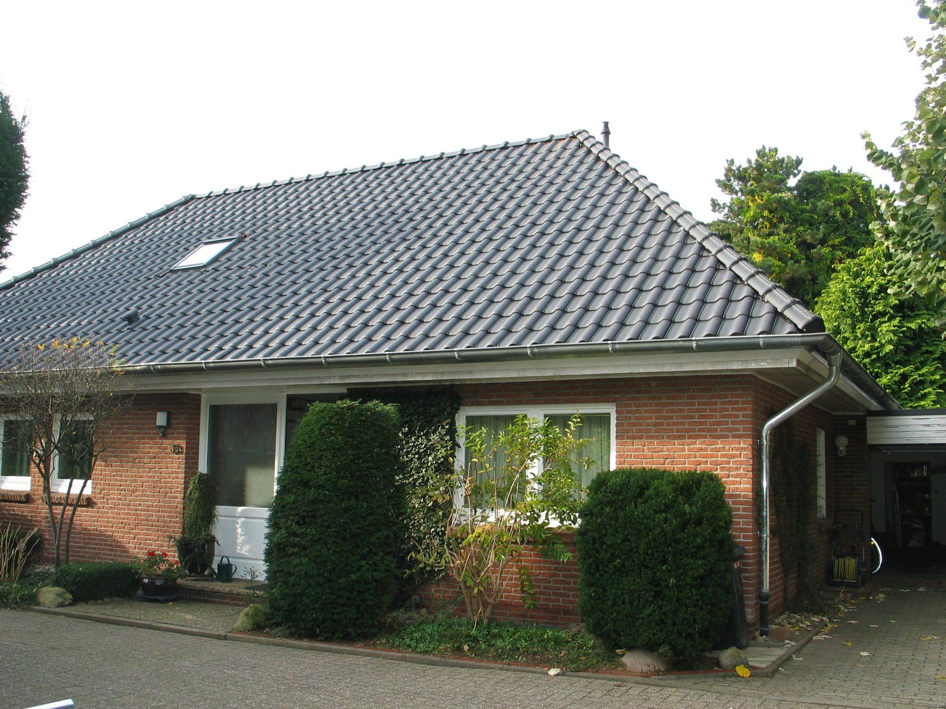 Rotes Backsteinhaus mit dunklem Ziegeldach, Bäumen und einem Carport.
