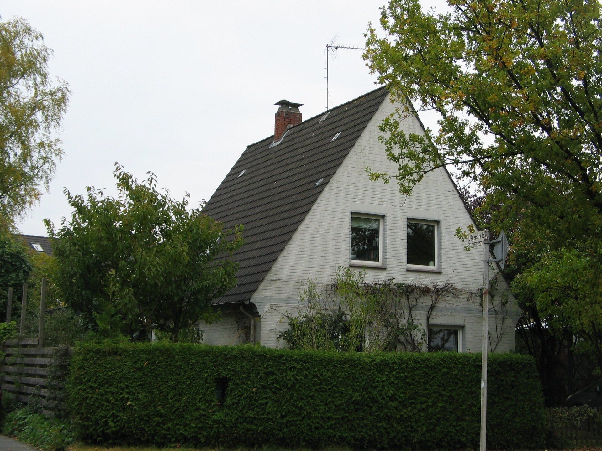 Weißes Haus mit dunkelgrauem Dach, zwei Fenstern, Schornstein, umgeben von grünen Bäumen und Sträuchern.