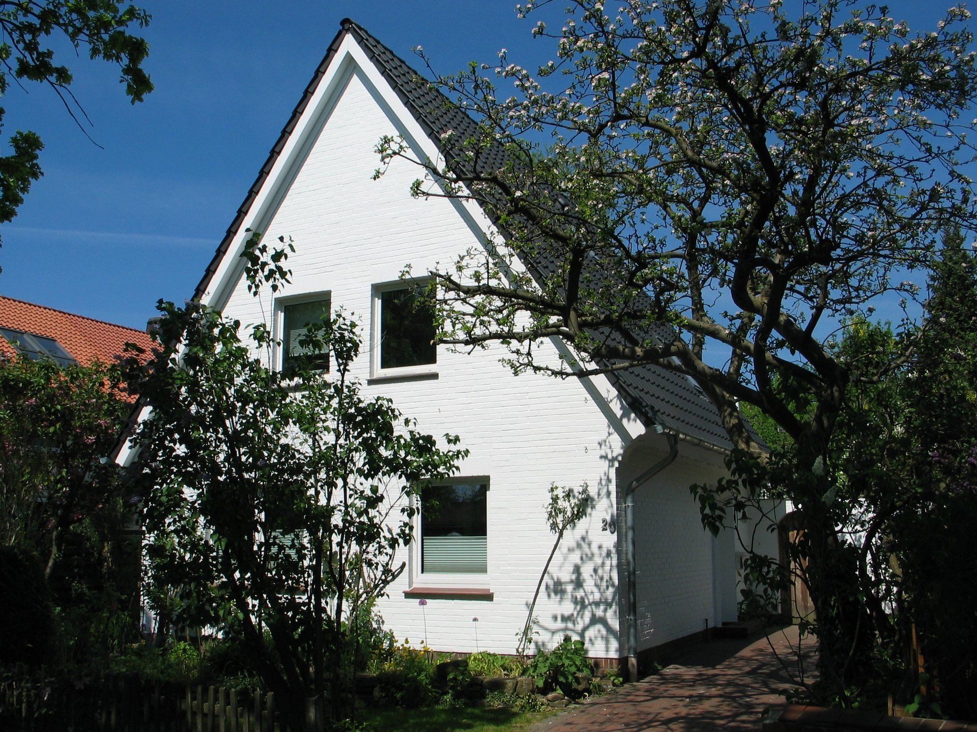 Weißes Haus mit schwarzem Dach, zwei Fenstern und umgeben von grünen Bäumen an einem sonnigen Tag.