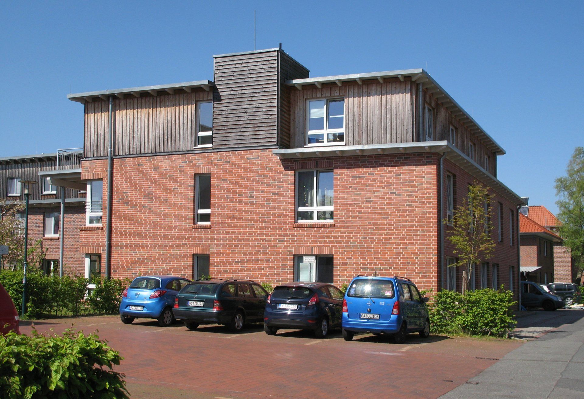 Rotes Backsteingebäude mit Holzelementen, davor geparkte Autos.