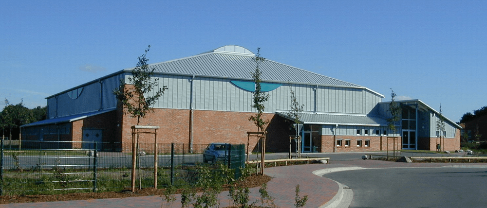 Großes Gebäude aus Ziegeln und Metall mit geschwungenem Dach. Davor ein kleiner Baum, an einem sonnigen Tag.