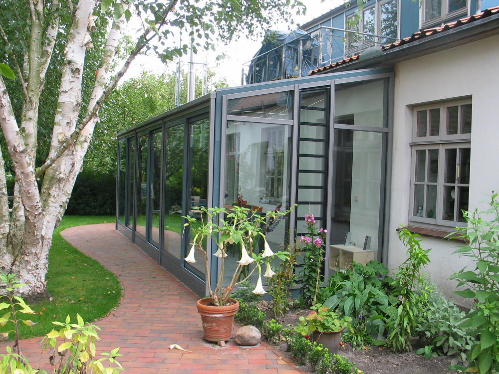 Ein verglaster Wintergarten, der an ein Gebäude angebaut ist, mit einem Ziegelweg, Topfpflanzen und einem Baum.