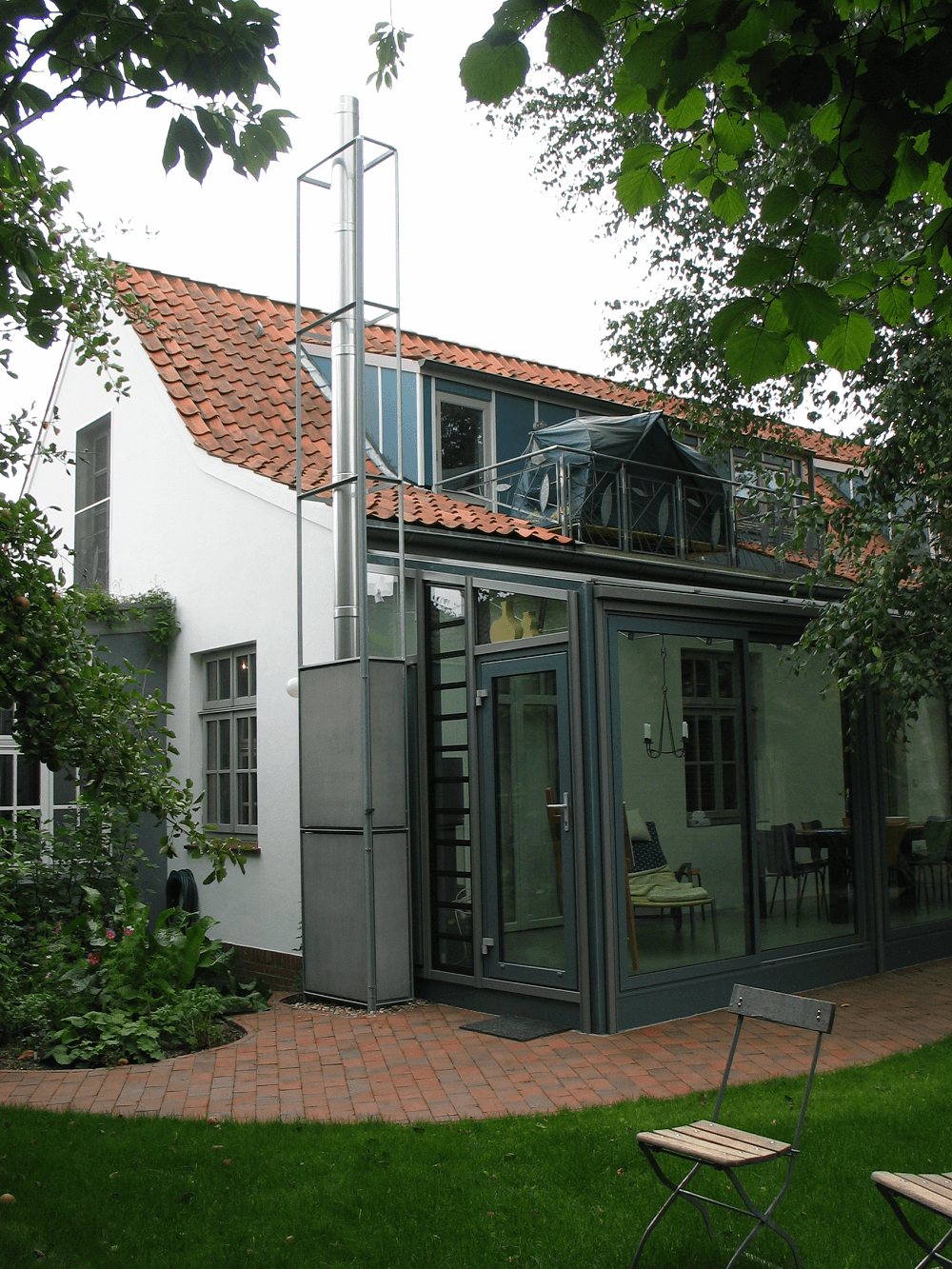 Weißes Haus mit rotem Ziegeldach, verglastem Anbau und hohem Schornstein, eingebettet in Grün und mit einer Ziegelterrasse.