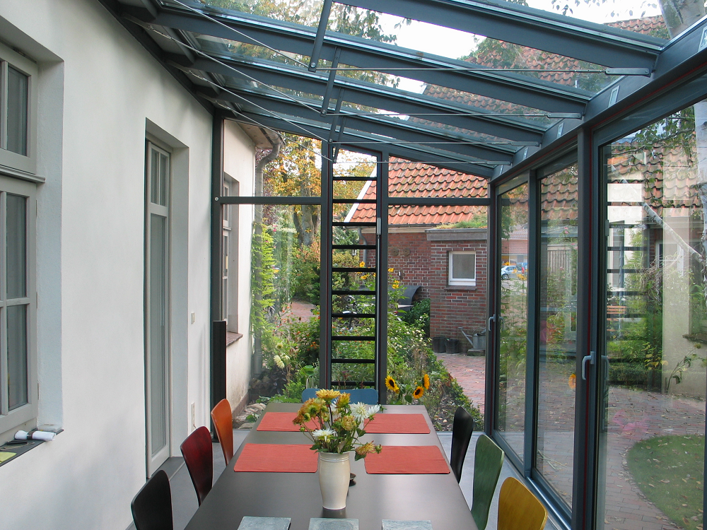 Wintergarten mit Glasdach und -wänden, ausgestattet mit Esstisch und Stühlen, mit Blick auf den Garten.