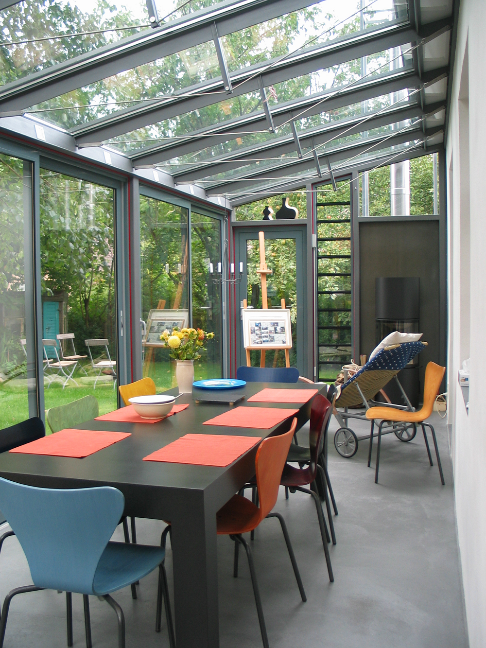 Wintergarten mit Glasdach und -wänden, Esstisch mit farbenfrohen Stühlen, Blick auf den Garten.