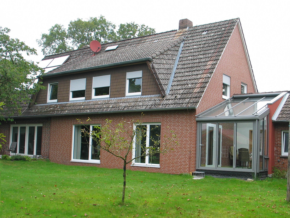 Backsteinhaus mit rotem Ziegeldach, Solaranlage und verglastem Seitenraum. Grüner Rasen davor.