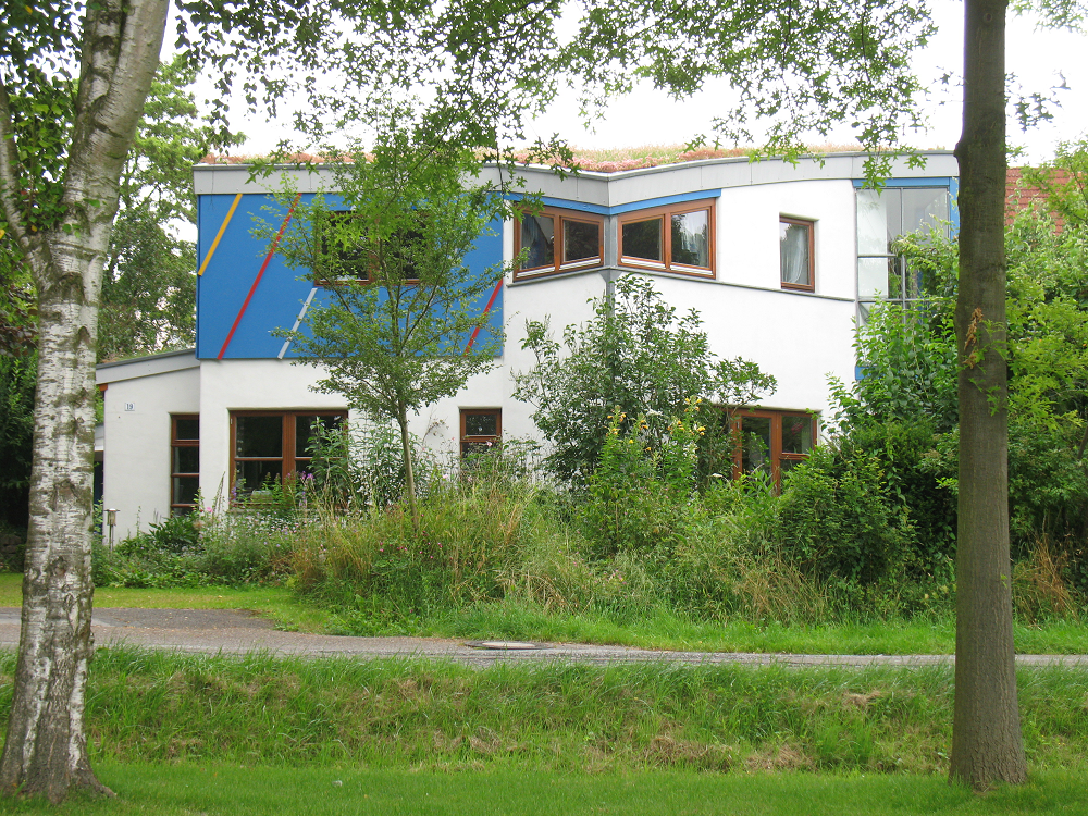 Weißes Gebäude mit blauen und gelben Akzenten, umgeben von Grün und Bäumen.