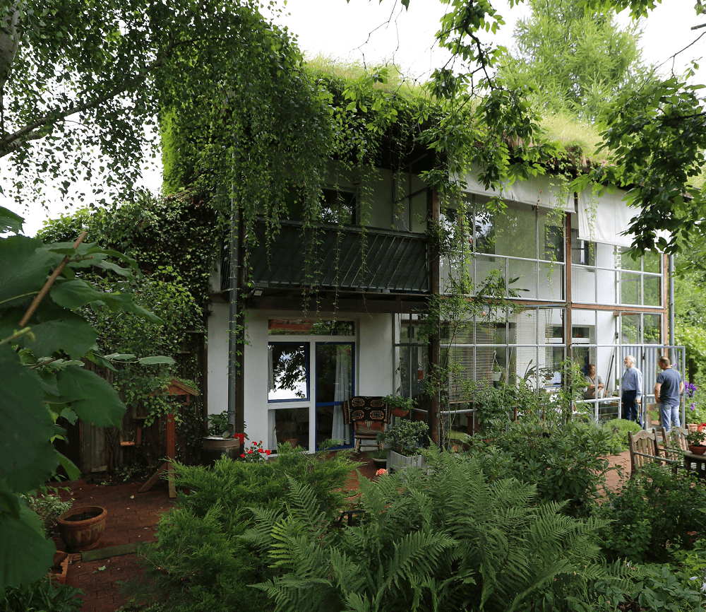 Weißes Haus mit großen Fenstern, Balkon und Weinreben, umgeben von üppigem Grün. Zwei Personen stehen daneben.
