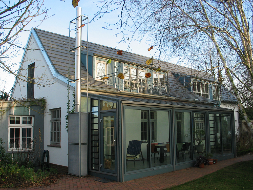 Weißes Haus mit verglastem Wintergartenanbau; Metallkamin; Bäume und Gras sichtbar.