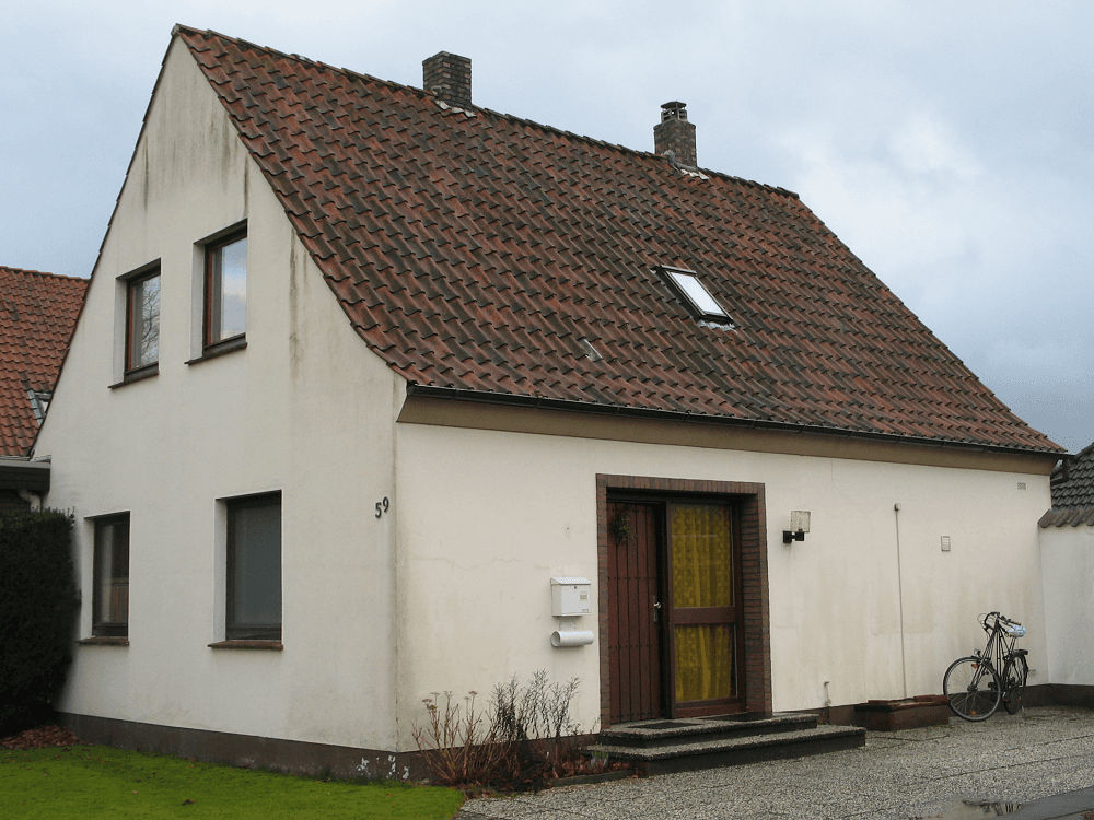 Beiges Haus mit rotem Ziegeldach, zwei Schornsteinen und einem Fahrrad, das in der Nähe auf einer Kiesauffahrt geparkt ist.