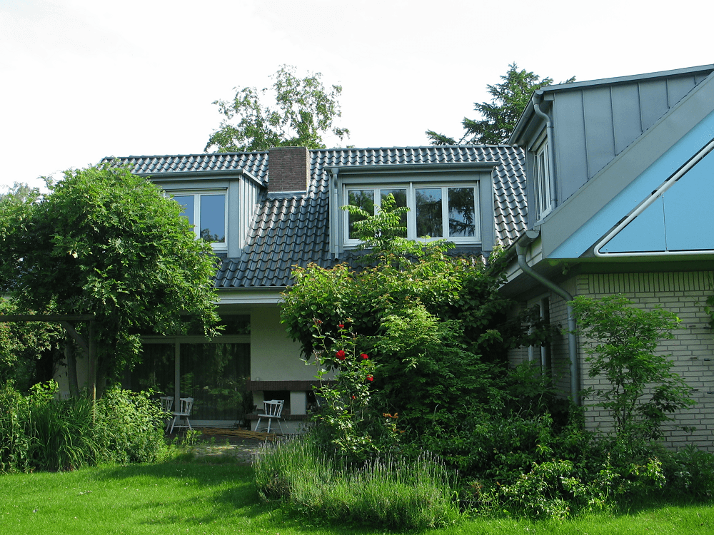 Haus mit dunkelgrauen Dachziegeln, Dachgauben und üppigem Grün im Vorgarten.