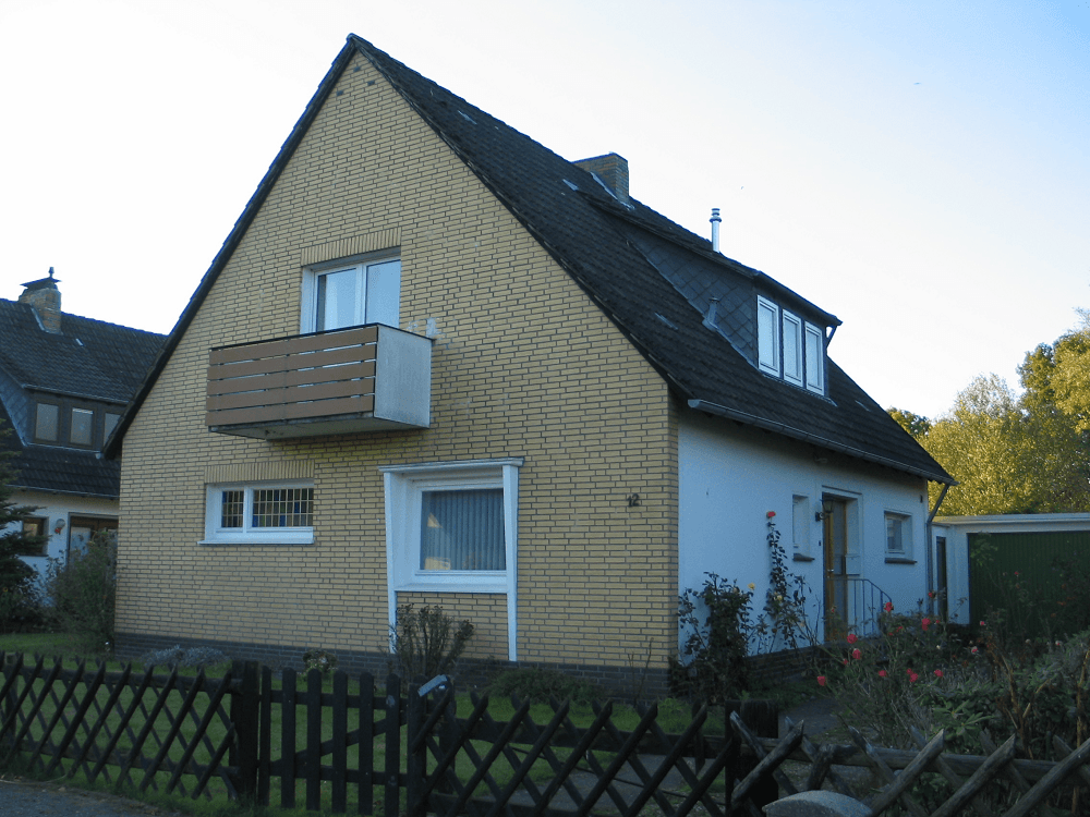 Haus mit hellgelber Fassade, weißen Wänden, schwarzem Dach, kleinem Balkon und dunklem Zaun.