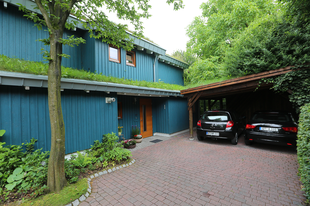 Blaues Haus mit grünem Dach und Carport, zwei Autos parken auf einer gepflasterten Auffahrt.