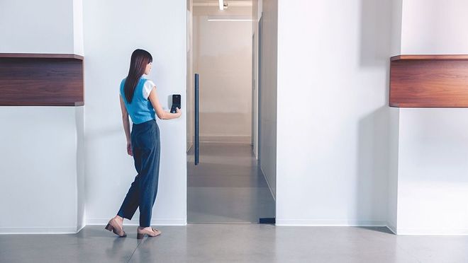 Woman in blue top unlocks door with a digital device. White walls and wooden shelves frame hallway.