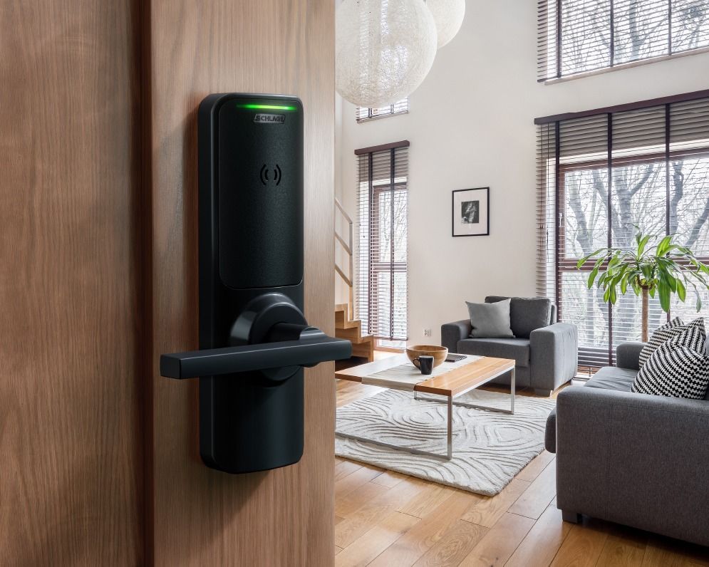 Black smart lock on a wooden door, open to a modern living room with gray furniture and natural light.