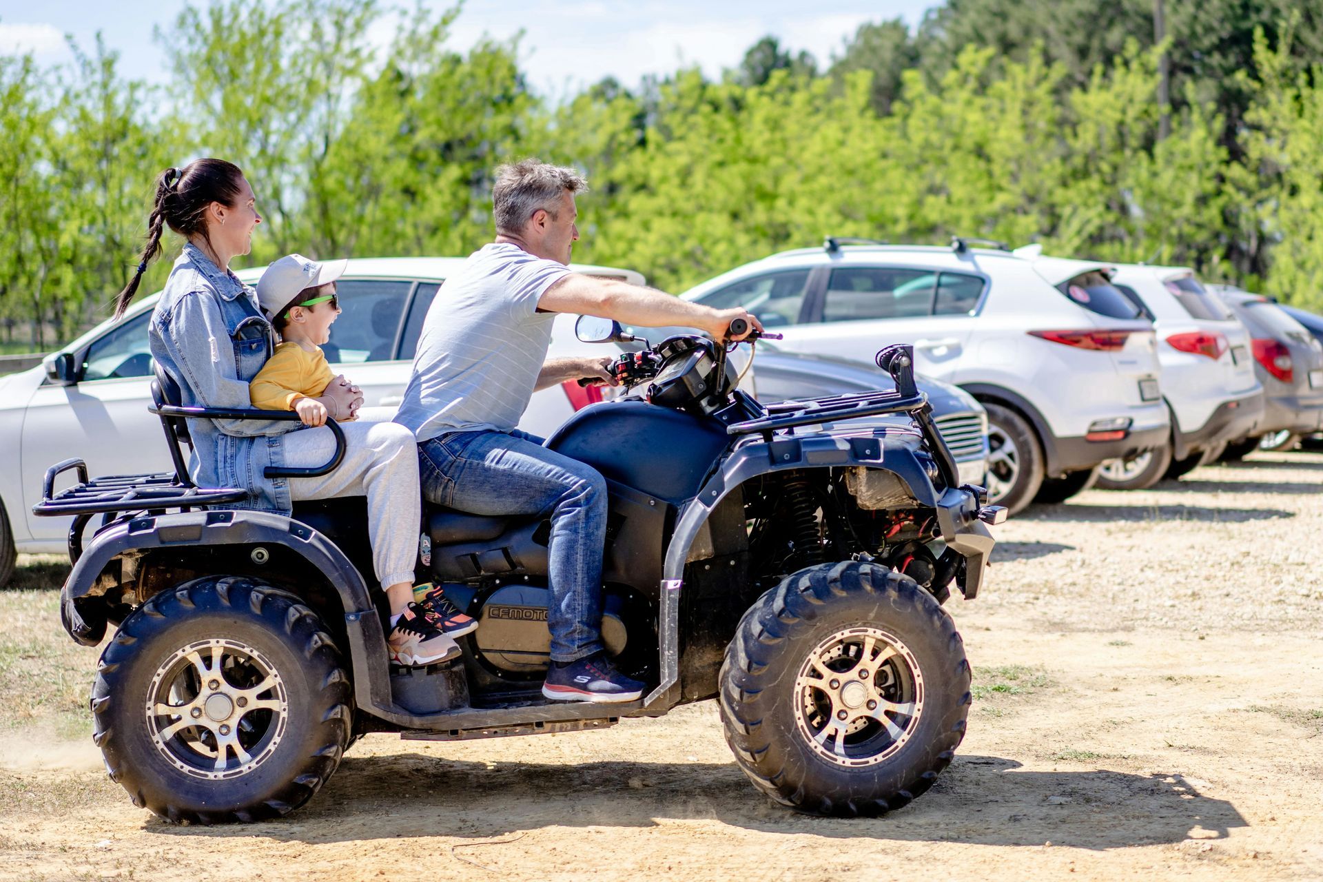 Family riding an ATV on a sunny day near parked cars and trees.