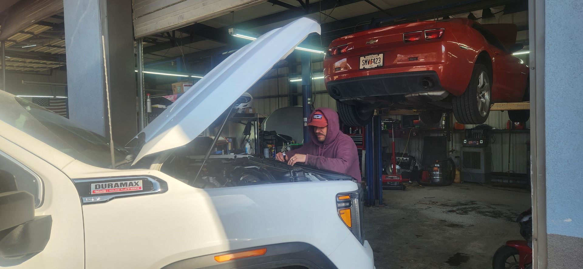 A mechanic works under the open hood of a white truck, with a red car hoisted on a lift in the background of a garage.