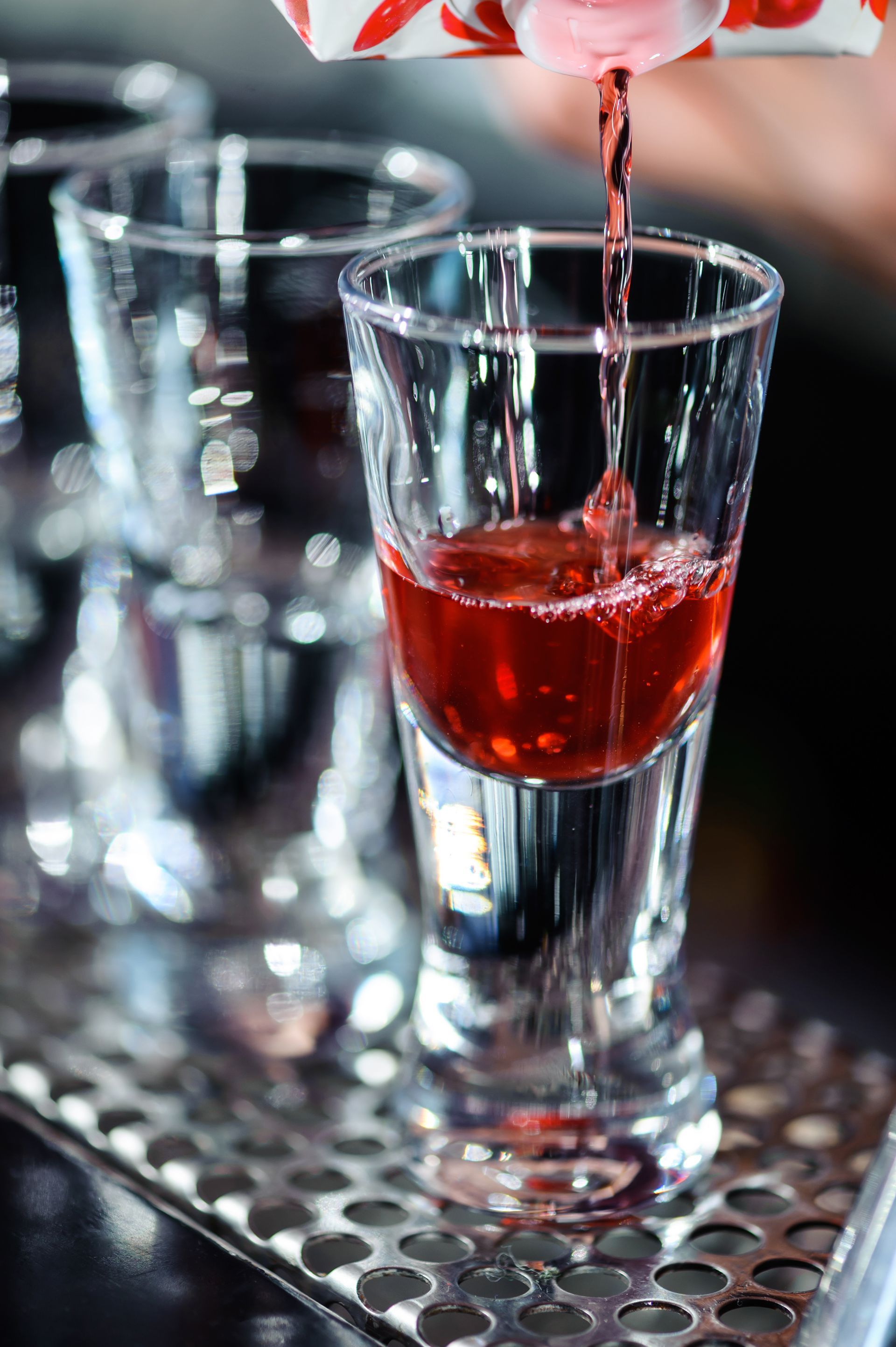 Red drink being poured into a clear shot glass on a reflective tray