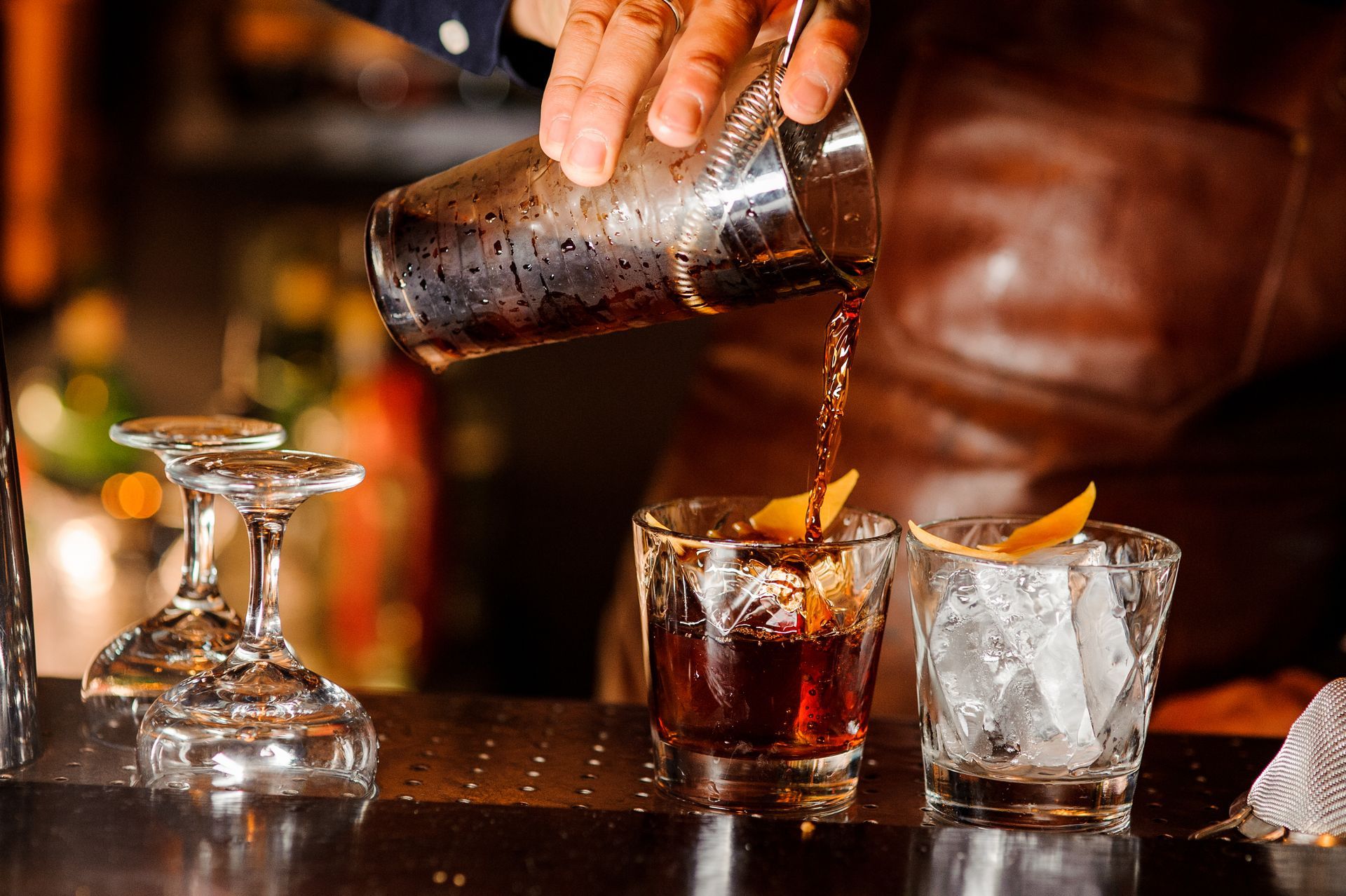 Bartender pouring a dark cocktail into a glass beside a whiskey tumbler with ice and orange peel.