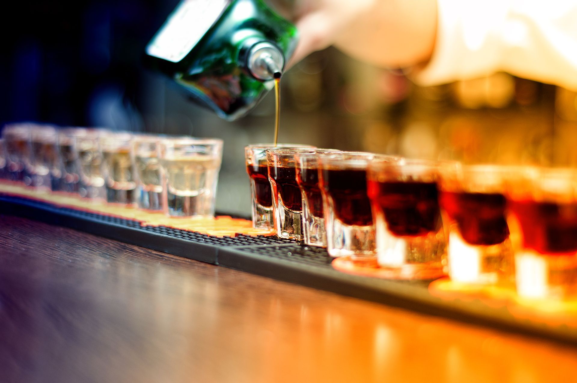 Bartender pouring shots of amber liquor into a row of clear glasses on a bar counter