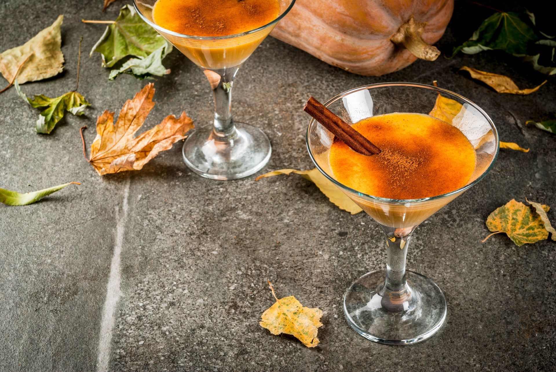 Two orange cocktails in martini glasses on a stone surface with autumn leaves and cinnamon sticks