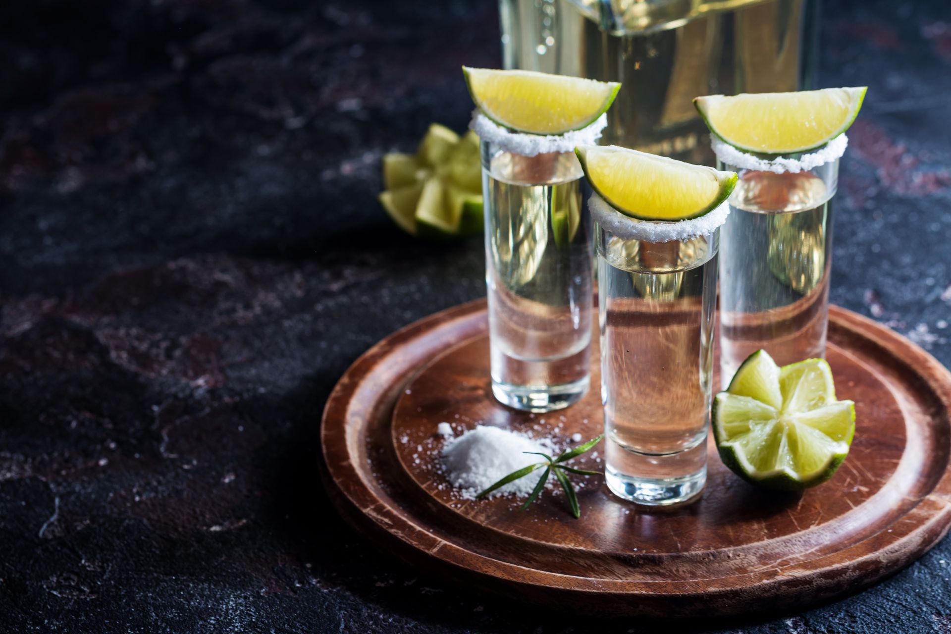 Three shot glasses of clear lime drink with lime wedges on a wooden tray, with a sliced lime nearby.