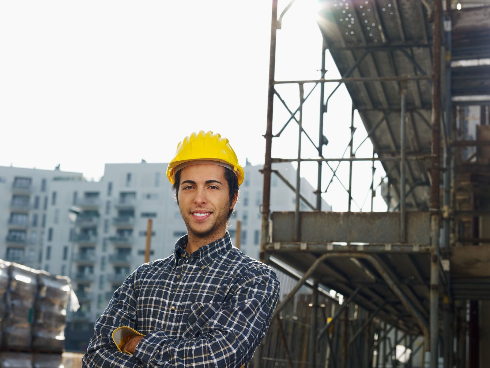 Construction worker wearing a yellow hard hat, smiling, arms crossed, in front of scaffolding and buildings.