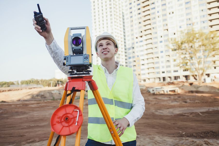 Construction worker using surveying equipment at a building site, holding a radio.