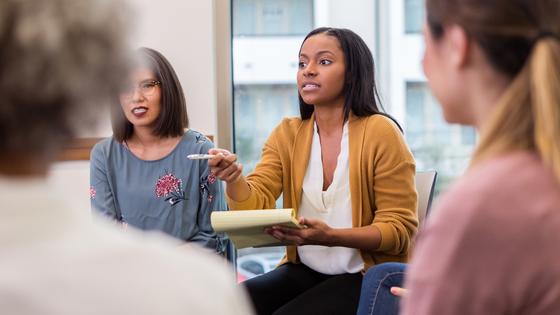 Woman points while speaking during a group discussion, holding a notepad.