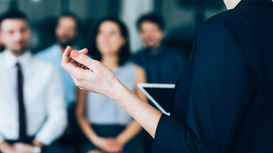 Woman presenting to a seated audience, gesturing with her hand.