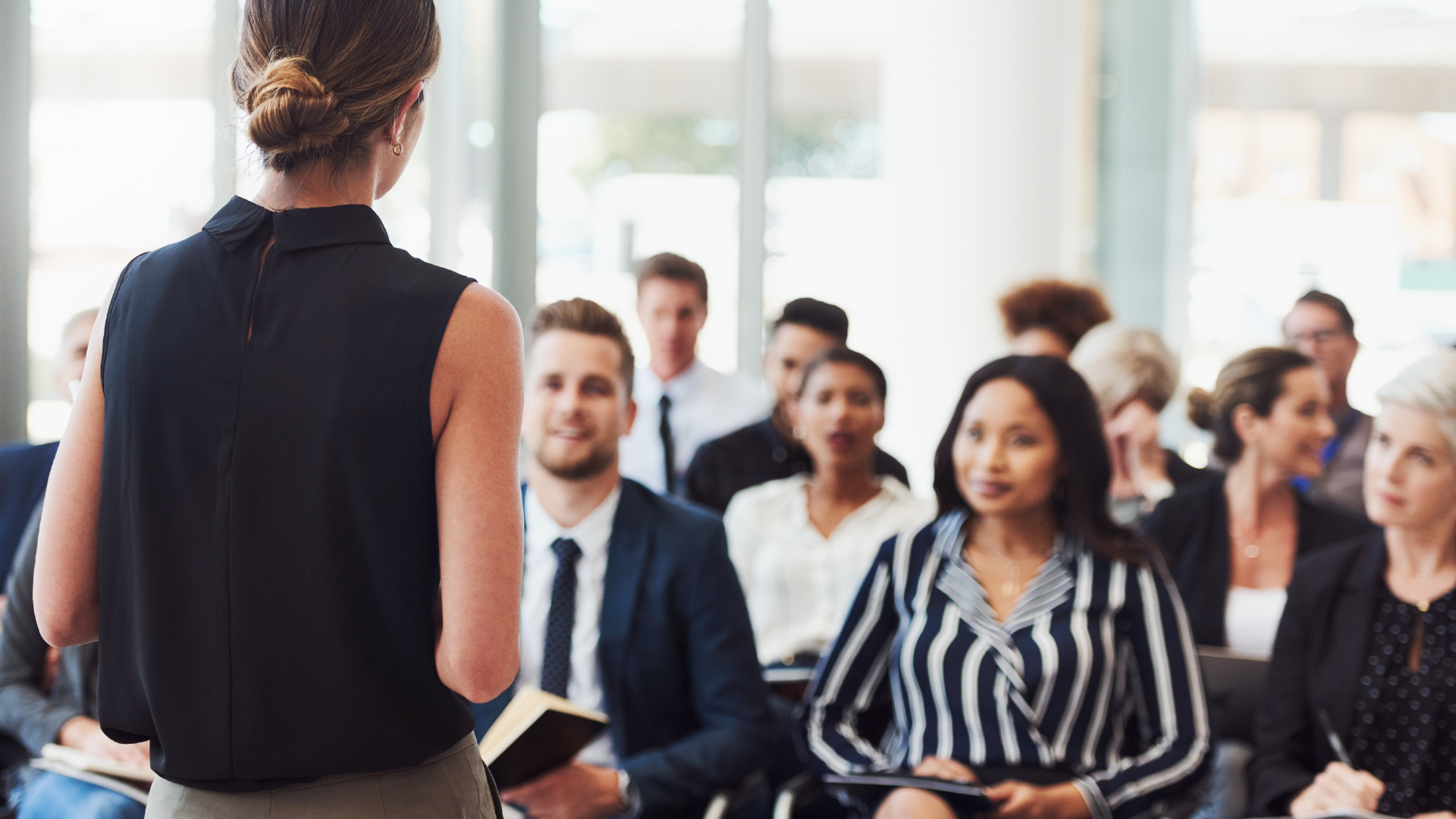 Woman speaking to audience in a modern conference room. People seated, attentive; some smiling.