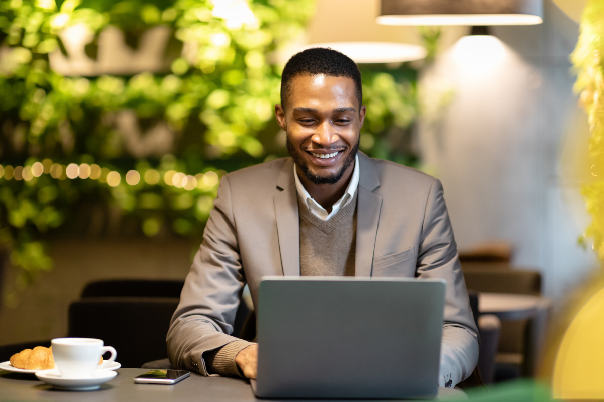 Smiling person in a blazer working on a laptop in a cafe with a coffee cup and greenery.