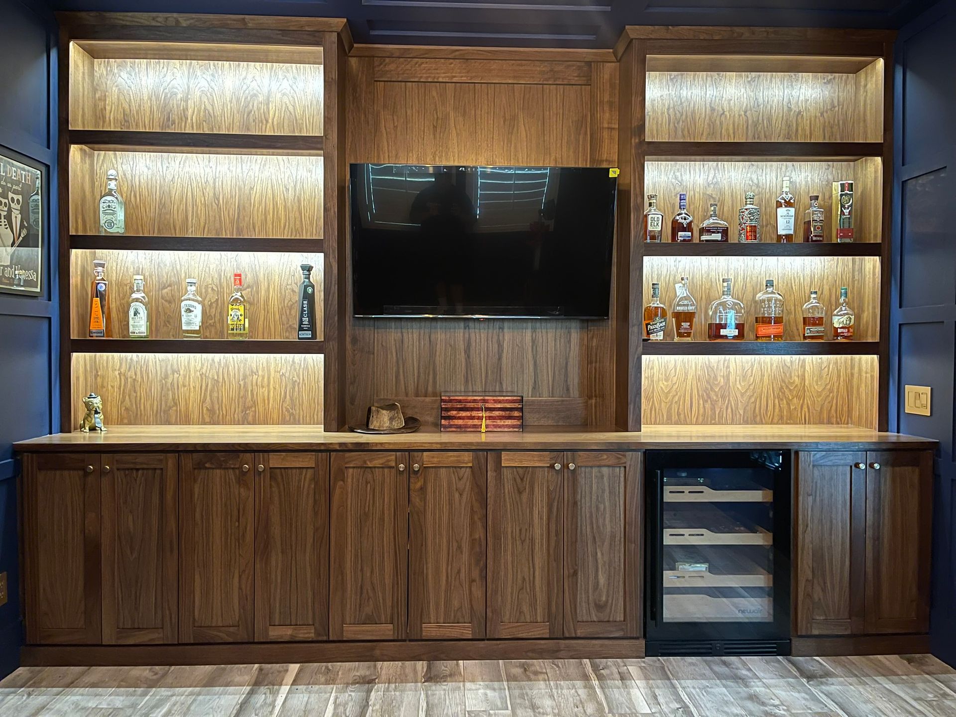 A home bar with wooden cabinets , shelves , a refrigerator and a flat screen tv.