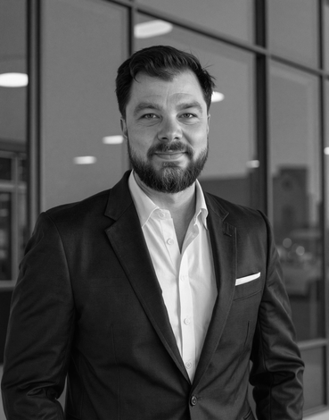 Man in a dark suit and white shirt, standing in front of a glass wall. He is smiling.
