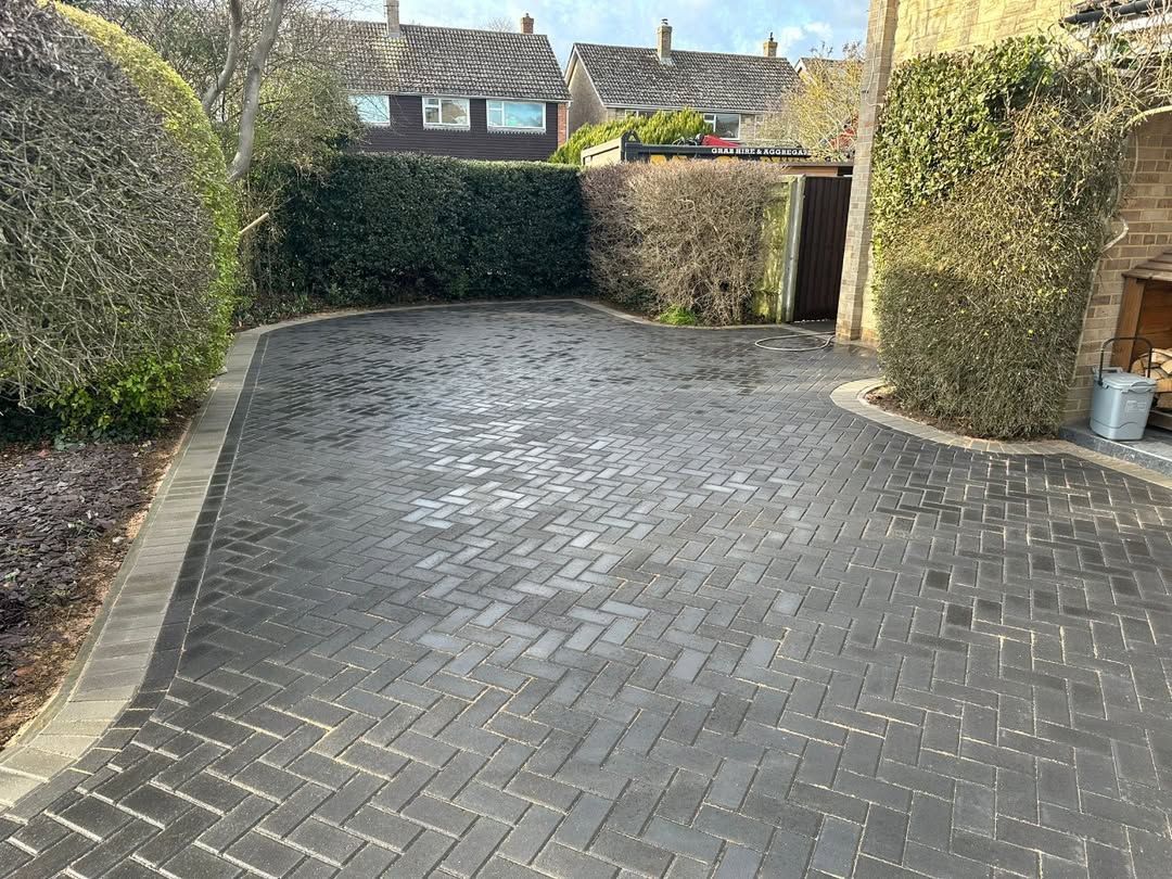 Brick-paved driveway bordered by hedges, leading to a closed gate and houses beyond