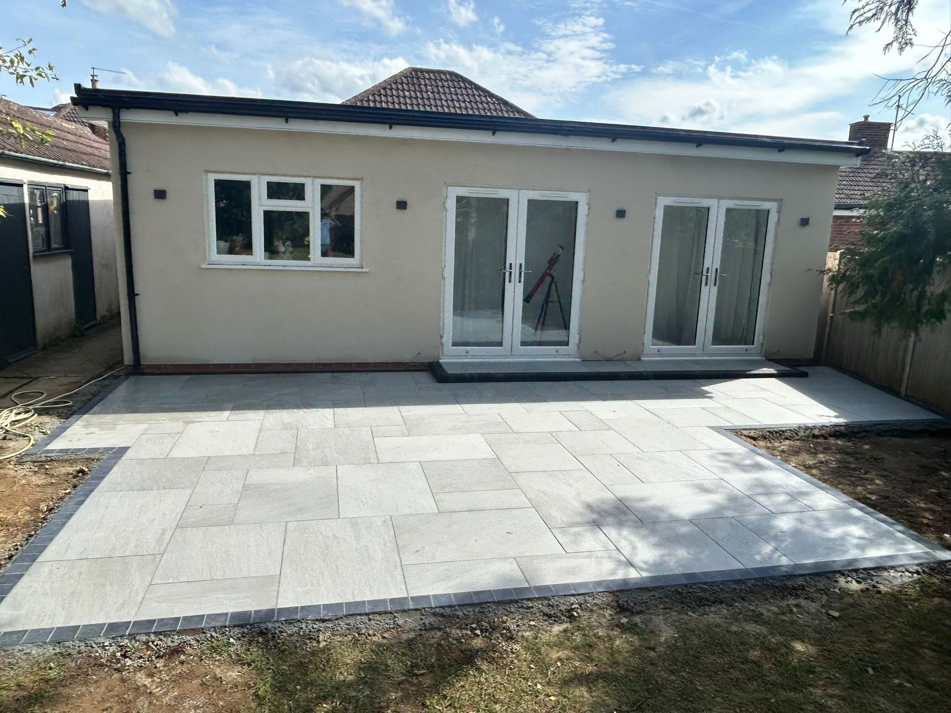 Backyard patio with light stone paving in front of a beige house with glass doors and windows