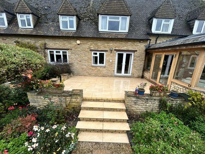 Stone house courtyard with steps leading to a patio, flower beds, and white-framed windows.