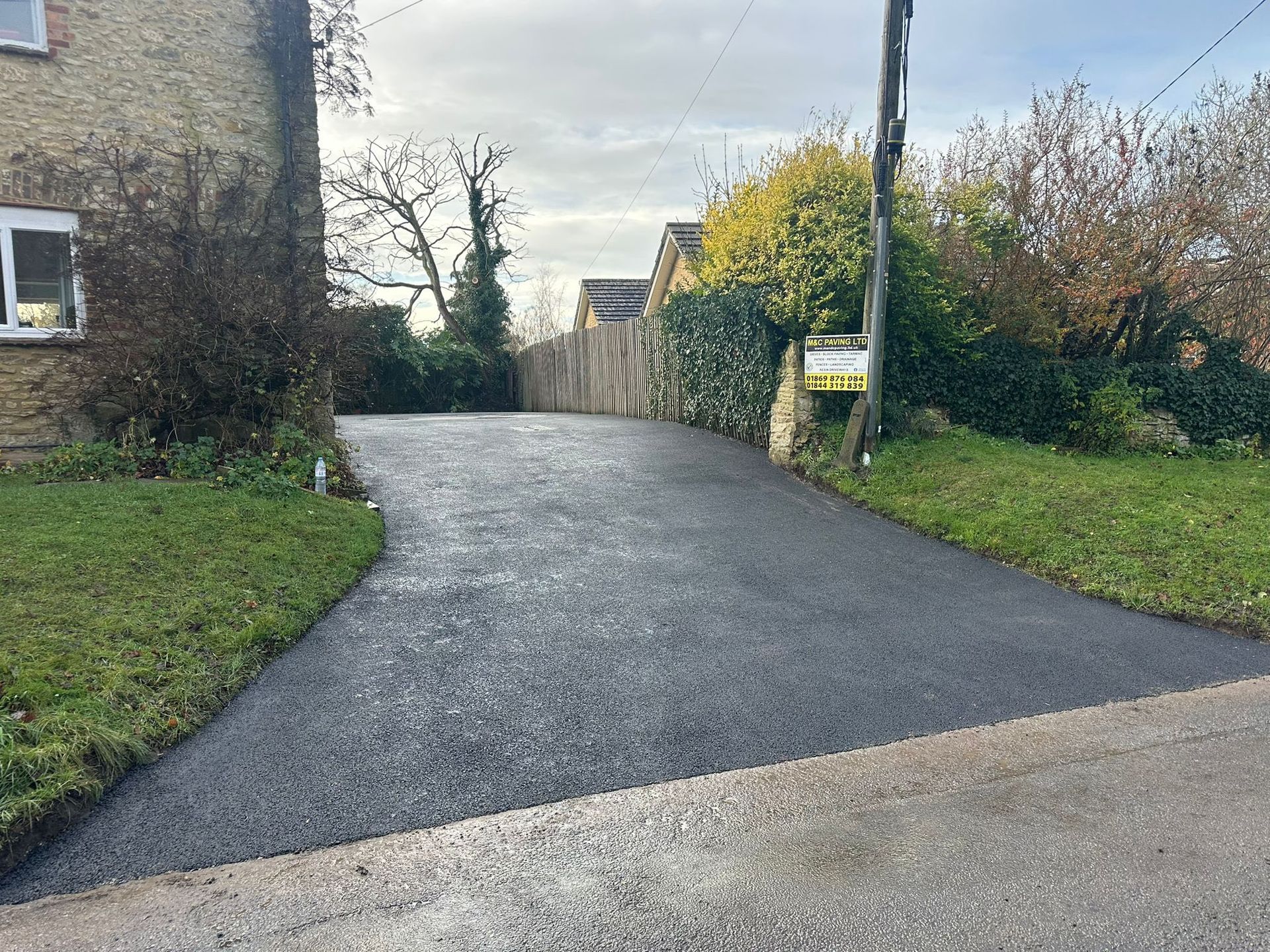 Freshly paved driveway curving uphill between stone wall and hedges under an overcast sky