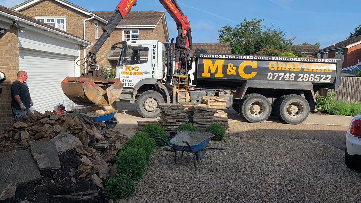 Construction truck unloading rubble in a residential driveway beside a house, with a worker nearby.