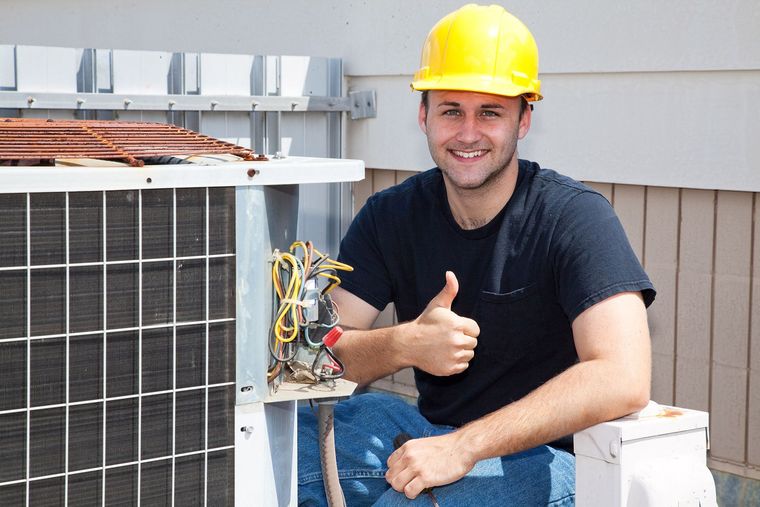 HVAC technician smiling and giving thumbs-up near air conditioner unit. Wearing a yellow hard hat and holding exposed wiring.