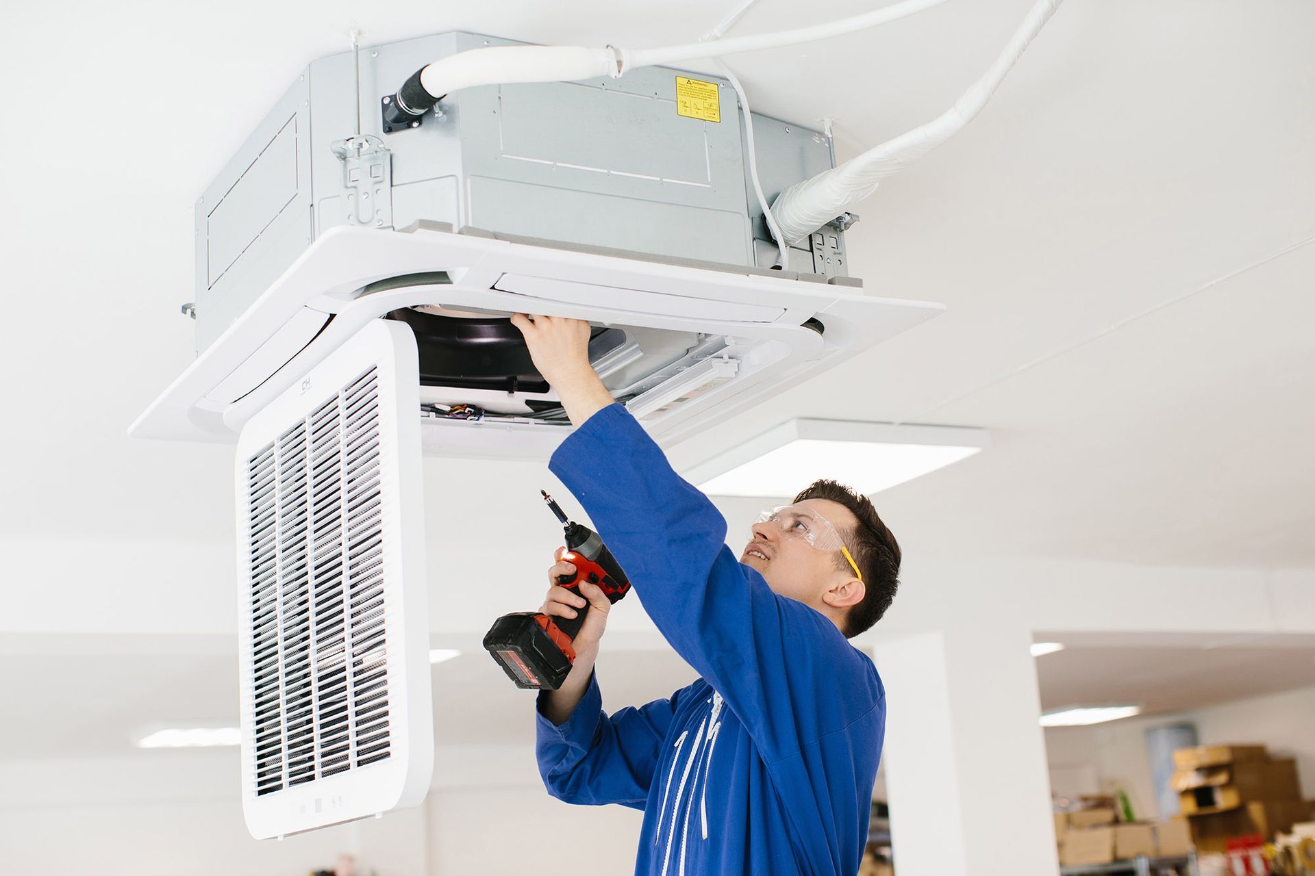 HVAC technician in blue overalls uses a drill to work on a ceiling-mounted air conditioning unit.