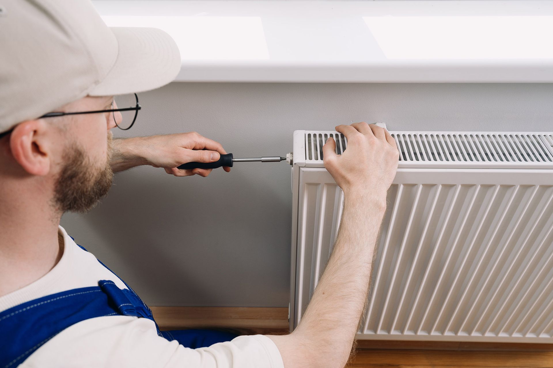 A person in overalls using a screwdriver to work on a white radiator.